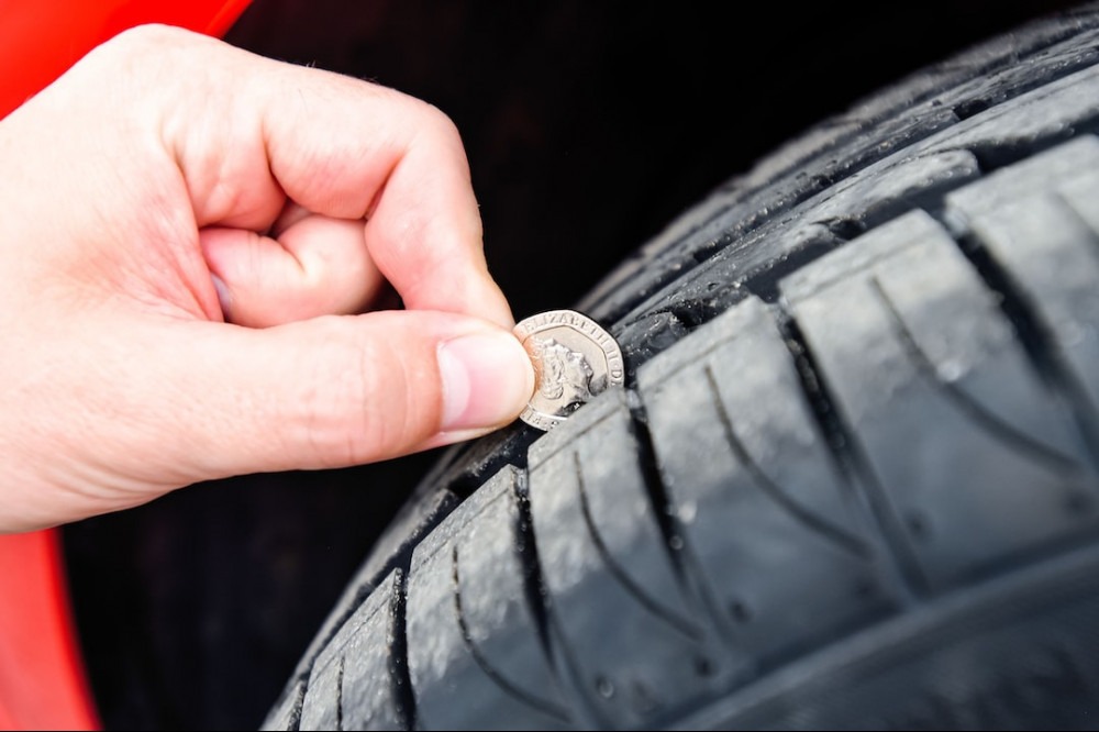 A hand of a person, holding a coin with the head of queen Elisabeth II, measuring the tread depth of a tire. The background is black, with a red detail behind the hand. He is following the car tire maintenance tips. 