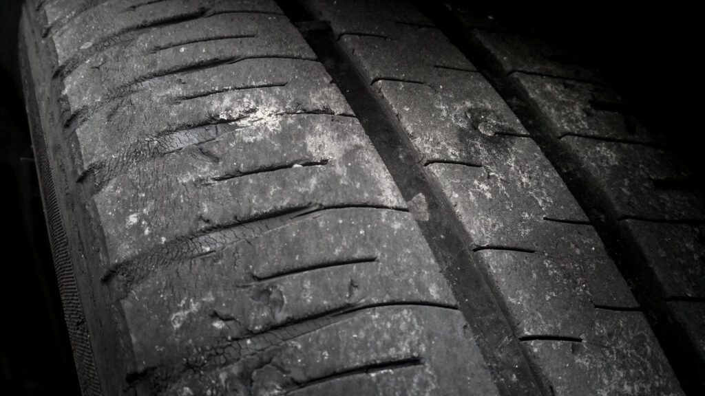 A close-up, low-angle shot of a severely worn-out car tire showing significant degradation of the tread. The tire is dark grey/black, and the once-defined grooves are shallow and uneven. There are visible cracks, chips, and areas where the rubber appears to be flaking or crumbling, particularly on the left side of the image. The texture of the tire surface is rough and pockmarked, indicating extensive use and wear. The background is dark, emphasizing the worn condition of the tire's surface.