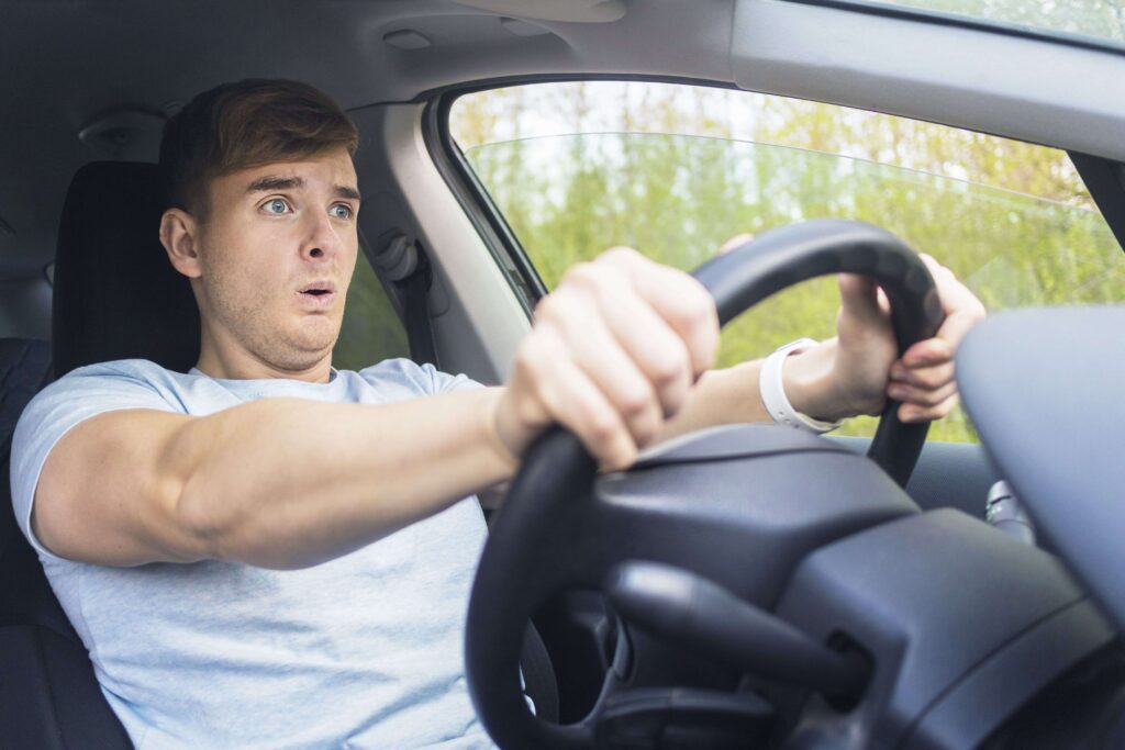 A young man with a concerned expression driving a car, suggesting the need for careful driving in wet conditions to avoid losing control.