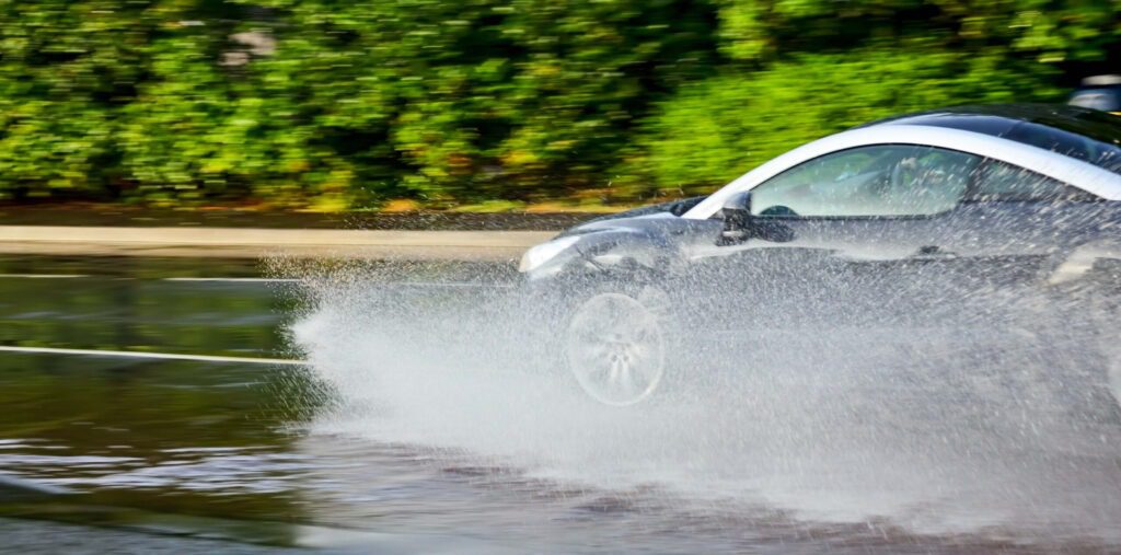 A car driving rapidly through a wet environment, with water spraying up around the wheels, representing the hazardous situation of hydroplaning where the vehicle's tires may lose contact with the road surface.