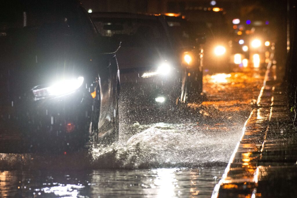 Cars driving through a flooded road at night, with water splashing up around the vehicles, indicating a high risk of hydroplaning.