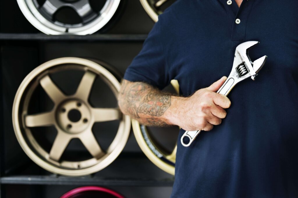 A man wearing a dark-blue t-shirt holding a wrench in his hand. The background is a bit blurry, but four rims can be seen behind the man. Three of them are goldish, and one of them is silver.