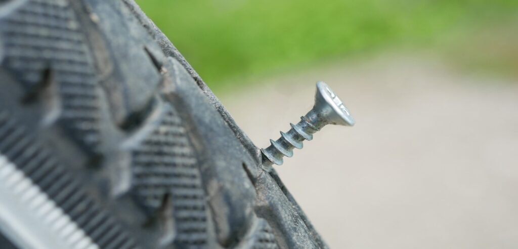 A zoomed-in car tire with a screw embedded inside. The background is blurry and the sole focus is on the puncture of the tire. Representing how a tire can be damaged. 
