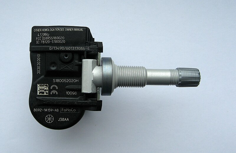 Close-up of a TPMS (Tire Pressure Monitoring System) sensor unit showing its internal components. The black plastic housing contains electronic parts and has a metal valve stem protruding from it. Part numbers and regulatory information are visible on the sensor body.