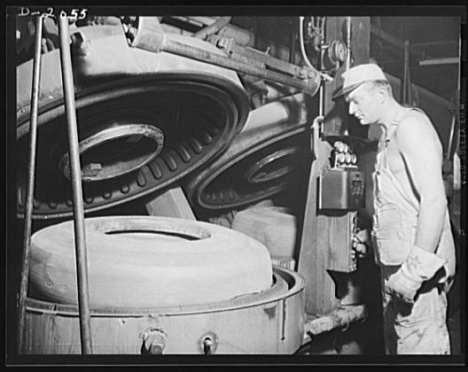 A black and white photograph from 1941 shows a factory worker overseeing a large, circular tire vulcanization mold. The worker, wearing light-colored overalls and a cap, stands to the right, looking intently into the machine's open cavity where a newly pressed tire sits. The heavy industrial machinery appears to be emitting steam or heat, characteristic of the tire manufacturing process of the era.