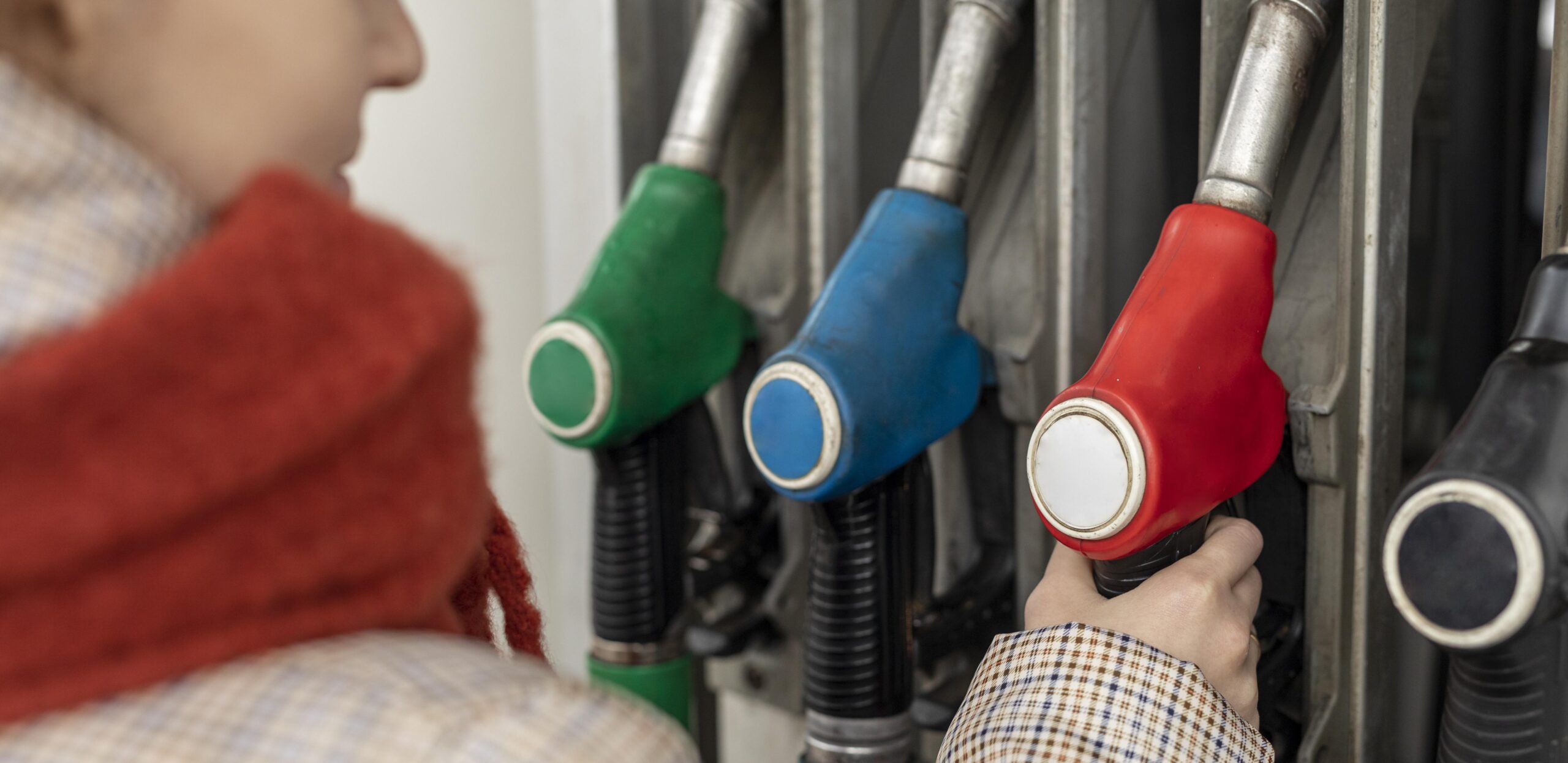 A woman at a gas station's fuel pump, reaching with her hand for the red fuel nozzle. Blurry background. The woman is dressed with a winter jacket.