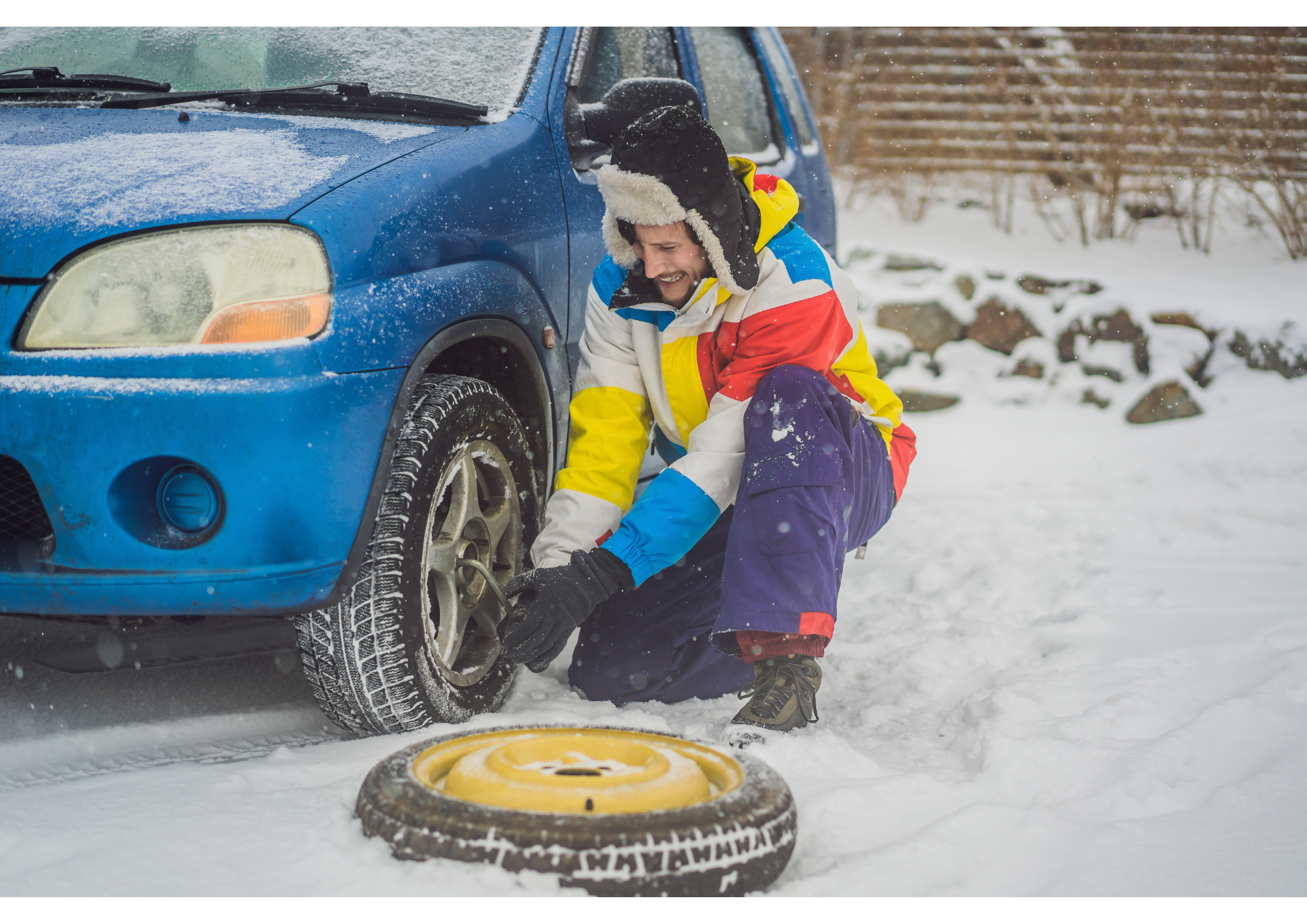 A man, well dressed for cold weather, changing the flat tire of his blue SUV. There is snow on the ground. At this moment, the man is loosening the wheel nuts, while the spare tire lays on the ground.