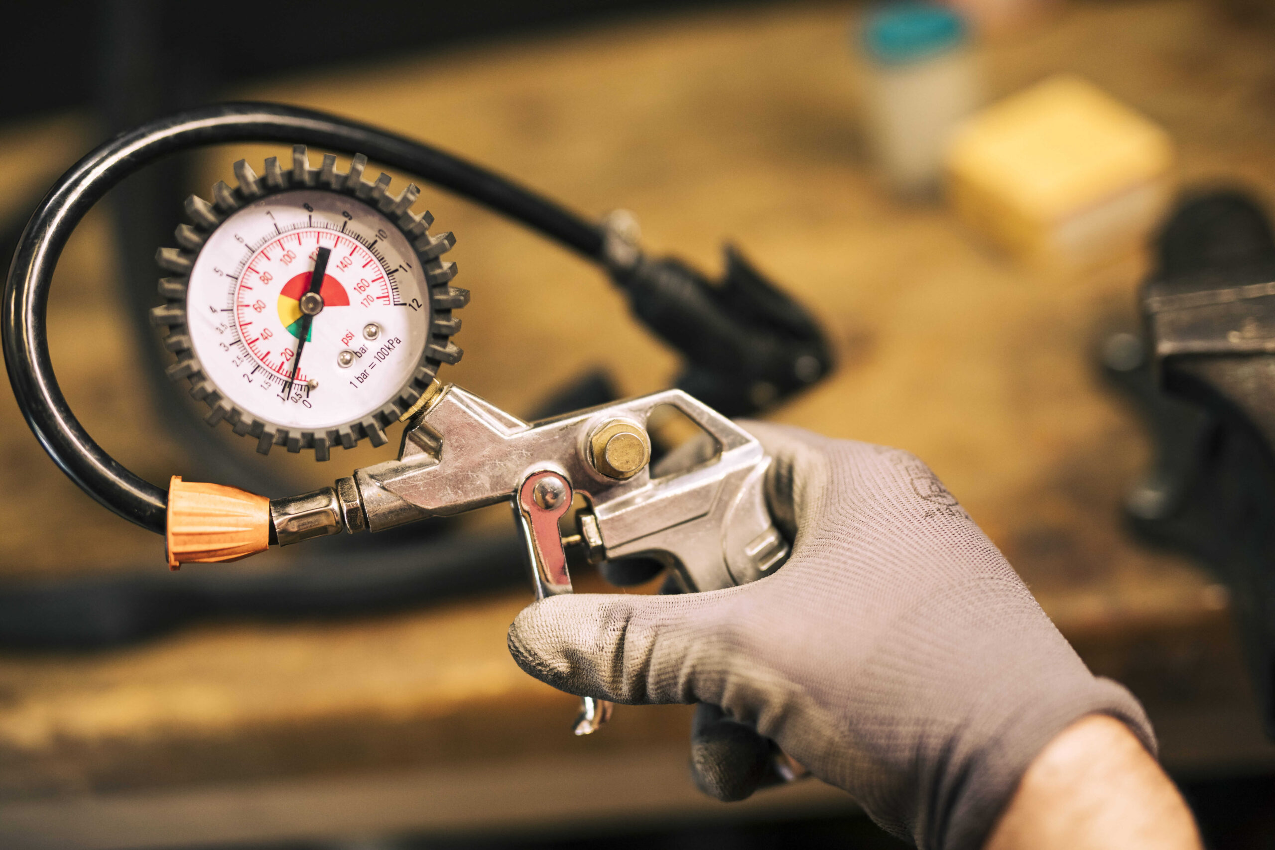 A close-up of a person's gloved hand holding a tire pressure gauge and an air pistol. The gauge is in sharp focus, showing the dial and hose. The blurry background of a cluttered workshop with a workbench and tools suggests the setting is a garage, relating to the process of checking for proper tire inflation.