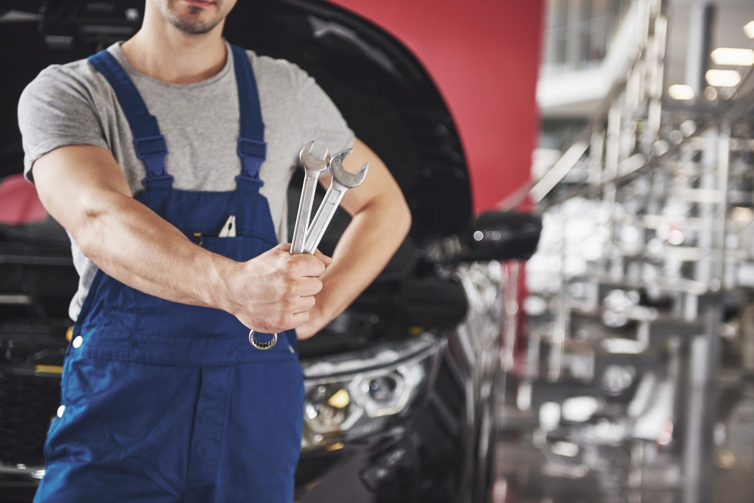 A mechanic in blue overalls holds two wrenches in front of a car with its hood open, symbolizing the importance of regular car maintenance.