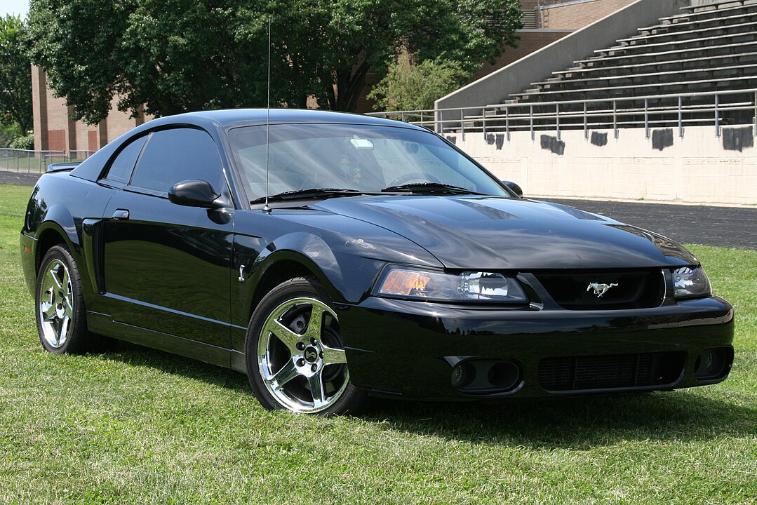 A 2004 Ford Mustang Cobra, part of the SVT lineup, captured in a close-up view of its front end. The car is finished in metallic black and features a more aggressive grille, distinctive hood vents, and Cobra badging, emphasizing its performance-oriented nature.