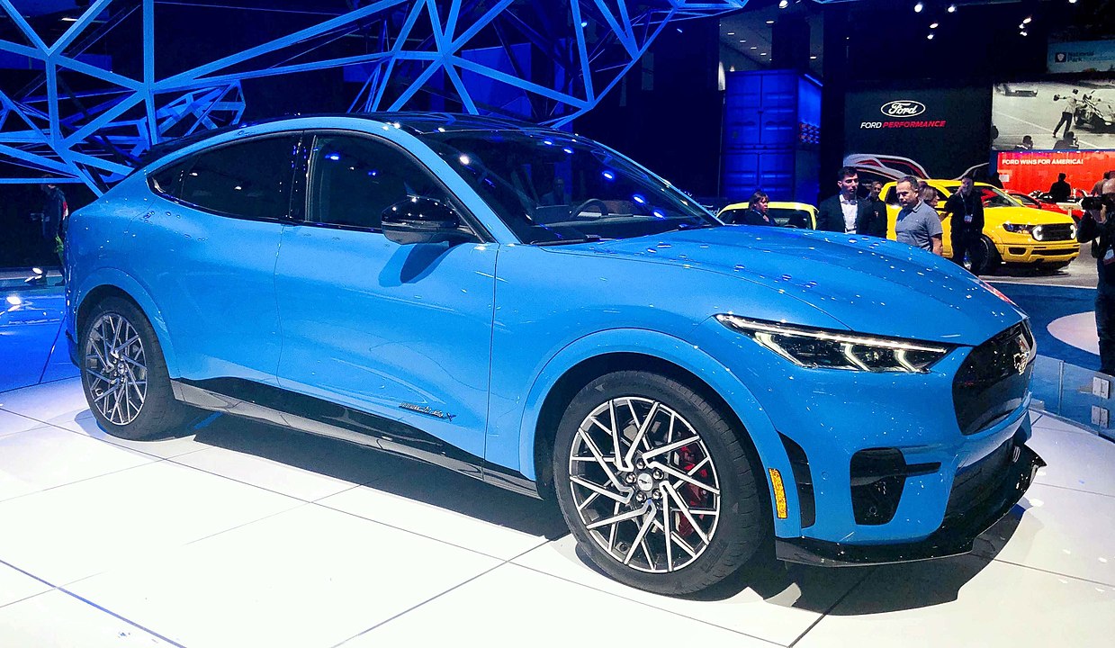 A Ford Mustang Mach-E GT, the electric performance SUV from Ford, finished in a vibrant blue color. The car features aerodynamic styling, a closed-off grille, sharp LED headlights, and bold black accents, redefining the Mustang nameplate in an all-electric era.