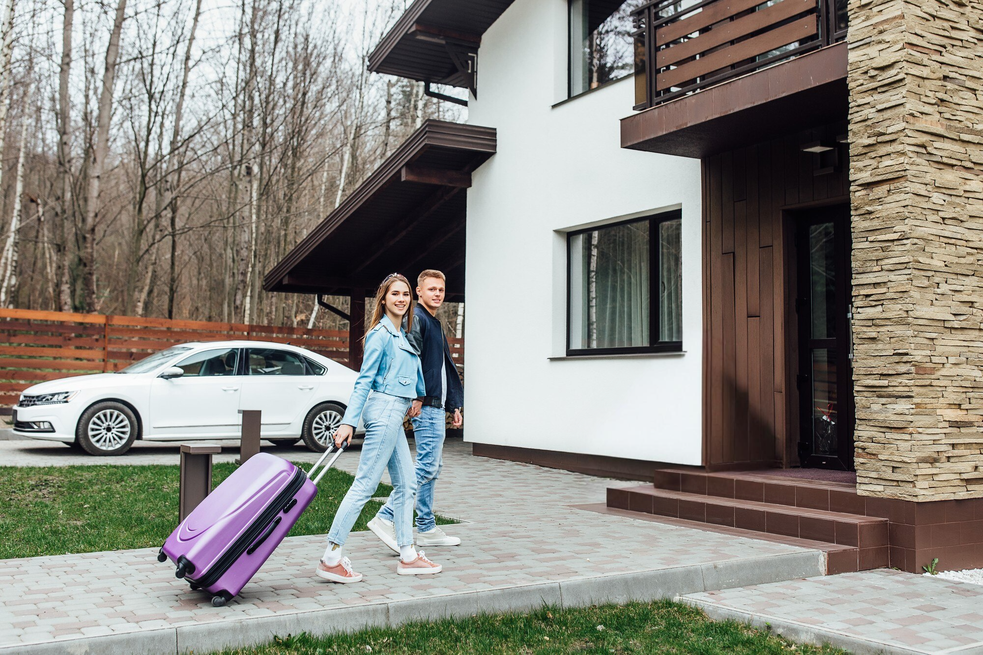 A couple arriving at a modern house with a white and stone exterior, the woman pulling a purple rolling suitcase while wearing a denim outfit. A white sedan is parked in the background, suggesting a road trip stop at their destination.