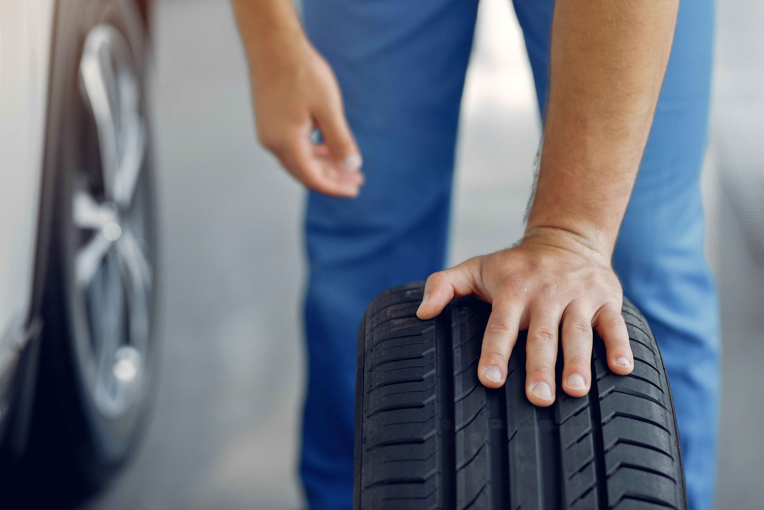 A man wearing blue working pants, holding a tire with one hand, while the other hand hangs freely in the air. There is a car next to him, with a focus on its tire, representing a man that is performing a tire rotation. 