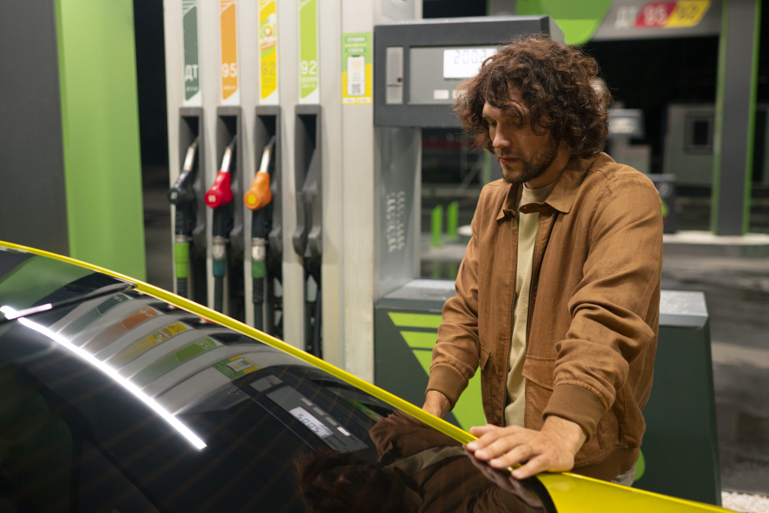 A man in a brown jacket at a modern gas station looking at his car, with multiple fuel pumps displaying different fuel grades visible in the background. The green accents and modern design of the station suggest it might be an eco-friendly or hybrid station offering both traditional and alternative fuels.