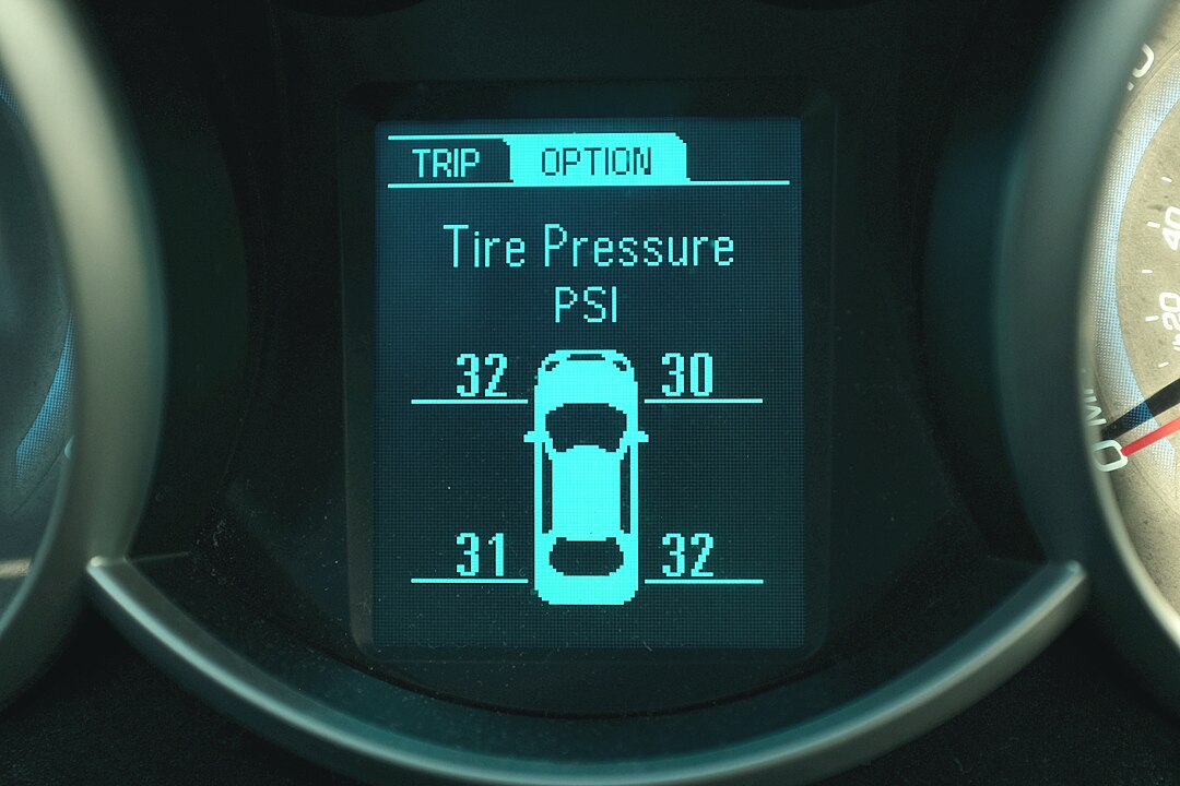 A close-up view of a vehicle's digital dashboard screen displaying the Tire Pressure Monitoring System (TPMS) readings. The screen is a bright cyan/blue on a dark background. The top of the screen shows "TRIP" and "OPTION" tabs, with OPTION currently selected. The main text reads "Tire Pressure" followed by "PSI." A stylized graphic of a car is centered, and four numbers indicate the pressure in pounds per square inch (PSI) for each tire: 32 (front left), 30 (front right), 31 (rear left), and 32 (rear right). The screen is framed by the dark gray plastic of the dashboard.