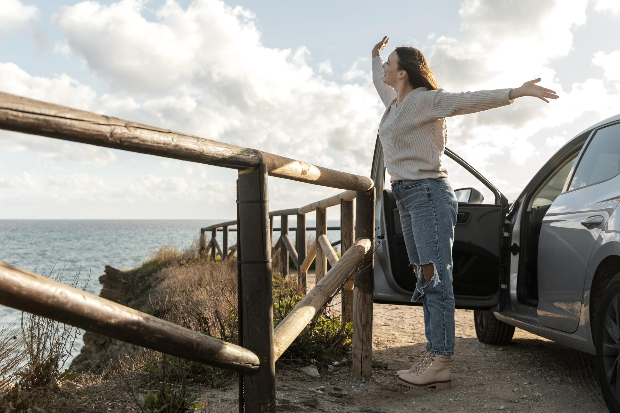 A woman standing with arms outstretched at a coastal viewpoint after arriving by car, demonstrating the importance of taking scenic breaks and stretching during long road trips.