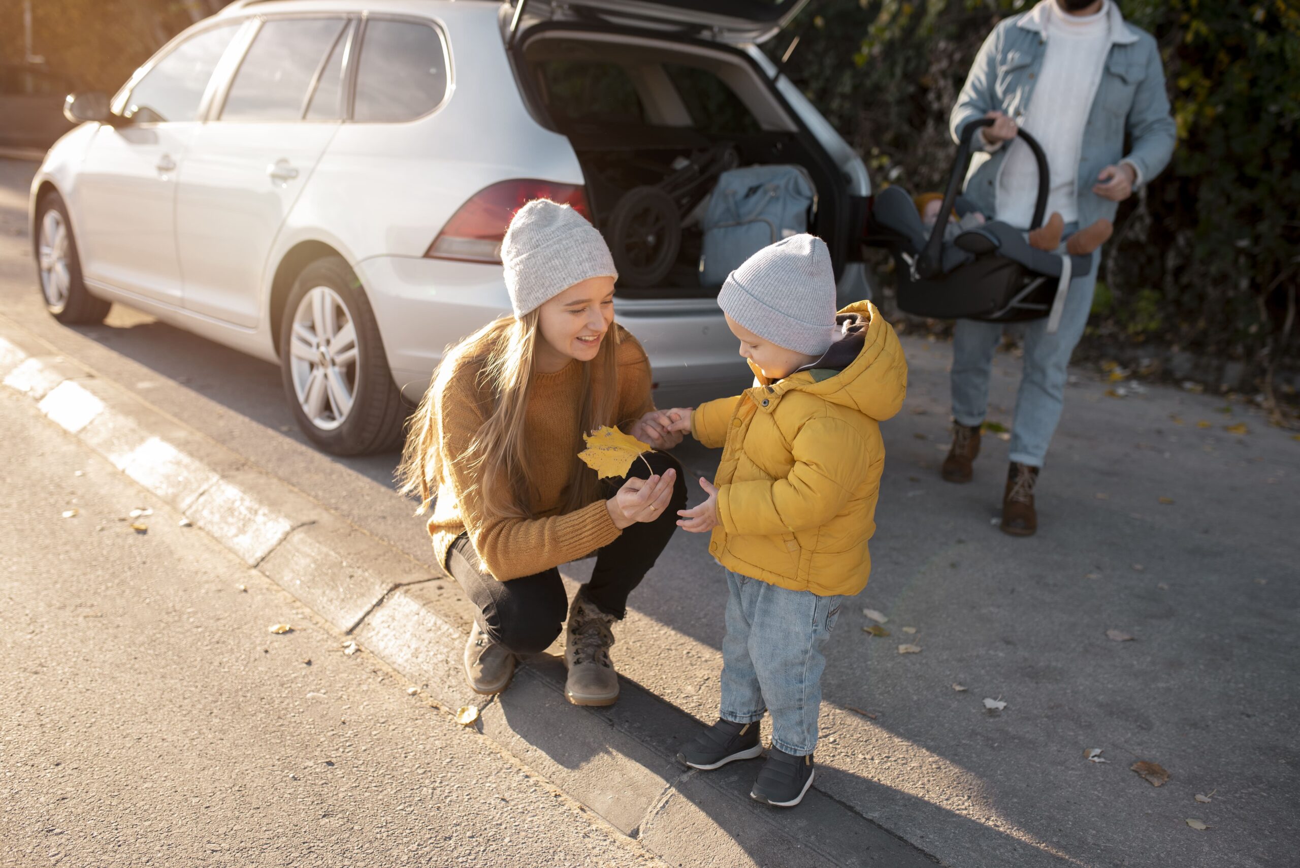 During a road trip break, a woman in a brown sweater and grey beanie crouches down to show a yellow autumn leaf to a small child in a yellow puffy jacket. A white SUV with an open trunk full of travel gear is visible in the background, highlighting the importance of taking breaks and engaging with surroundings during long drives.