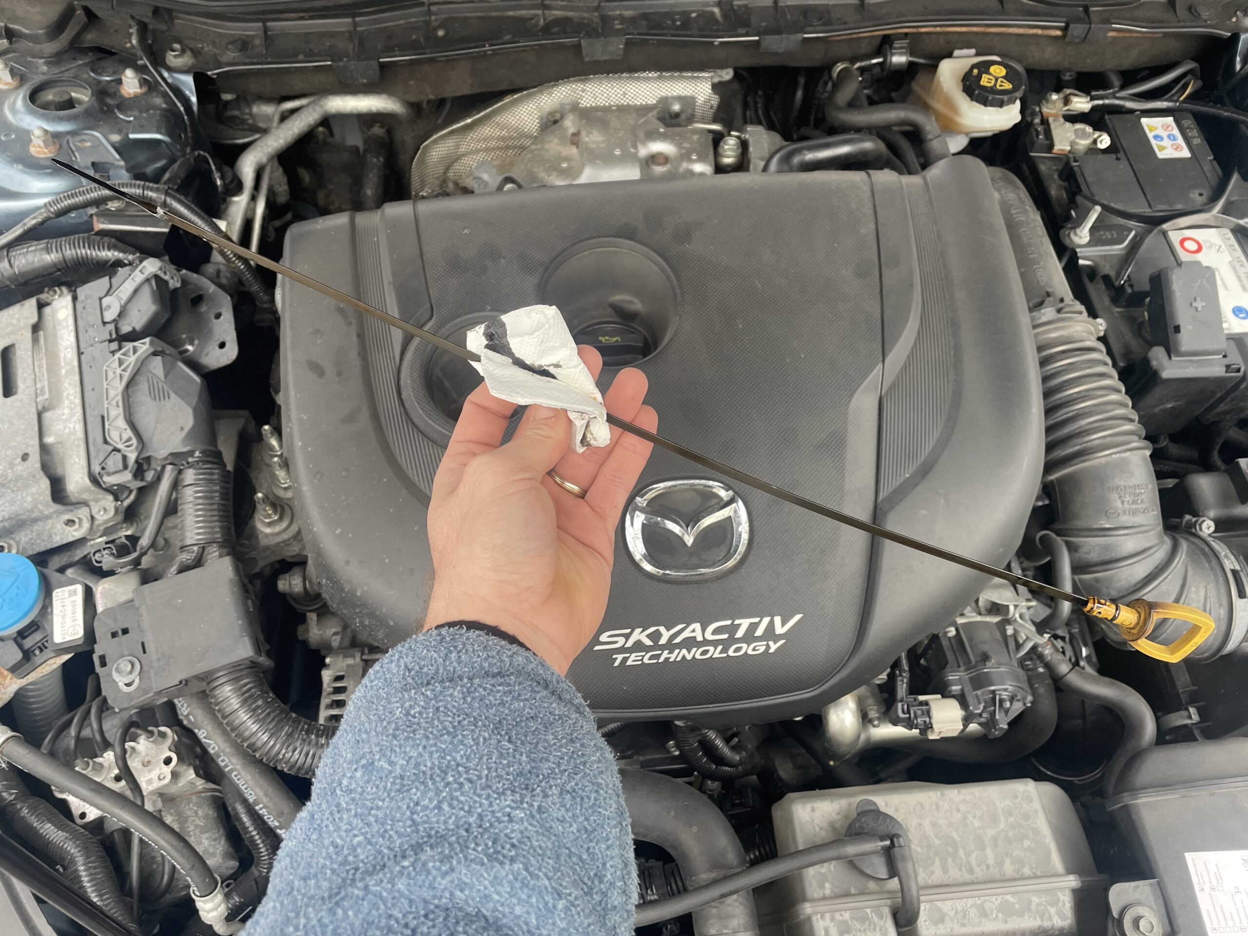 A hand holding the oil dipstick of a car. The ensigns and other parts of the car can be seen in the background. There is a huge logo of Mazda on the engine’s lid and the text “Skyactiv Technology”.