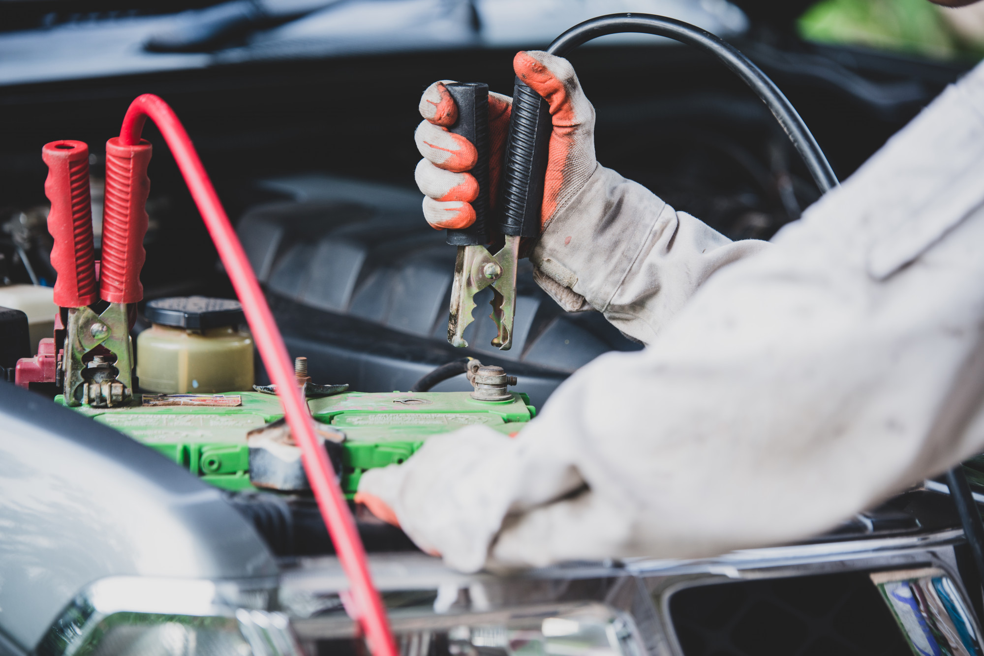 An image of a person's hands using jumper cables to connect a car's battery to another car's battery. The person is wearing protective work gloves and is carefully connecting the red and black clamps to the respective positive and negative terminals on the car's battery.
