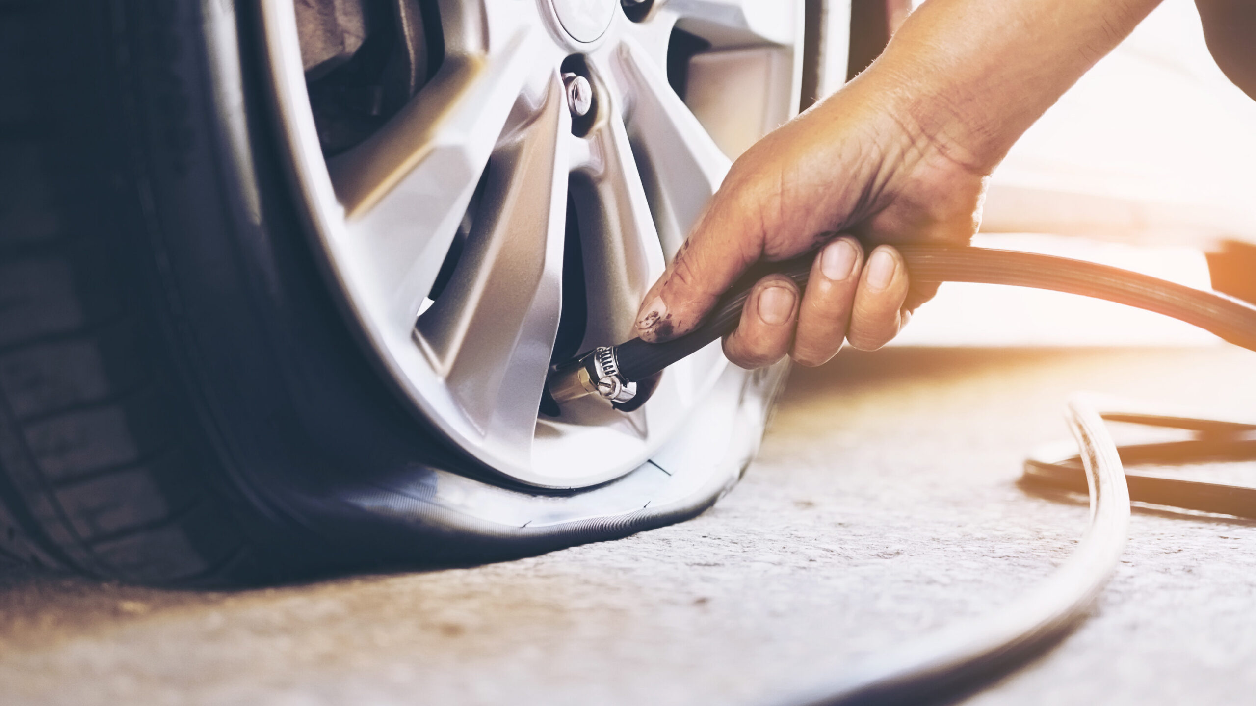 A close-up of a mechanic's hand inflating a car tire with a tire inflator.  The tire appears low on pressure, almost flat, as the mechanic connects the air hose to the valve stem. The image captures the importance of proper tire maintenance and addressing low tire pressure to ensure safe driving. Bright sunlight in the background adds a warm tone, highlighting the urgency of checking and inflating tires regularly. 
