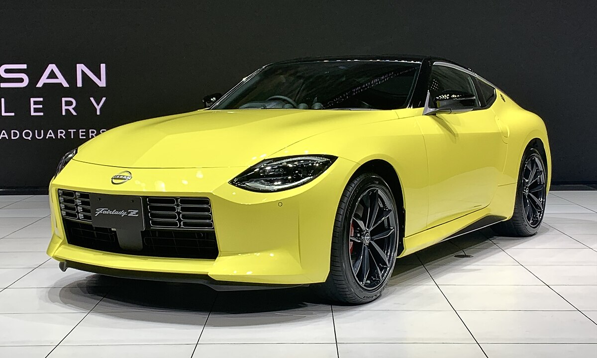 Front view of a bright yellow 2022 Nissan Fairlady Z (Z34) sports coupe displayed indoors at the Nissan Gallery Headquarters. The car features a retro-inspired design with sharp LED headlights, a wide blacked-out grille, and sporty black alloy wheels. This modern Z pays tribute to the classic Z lineage while incorporating contemporary styling cues and performance engineering.