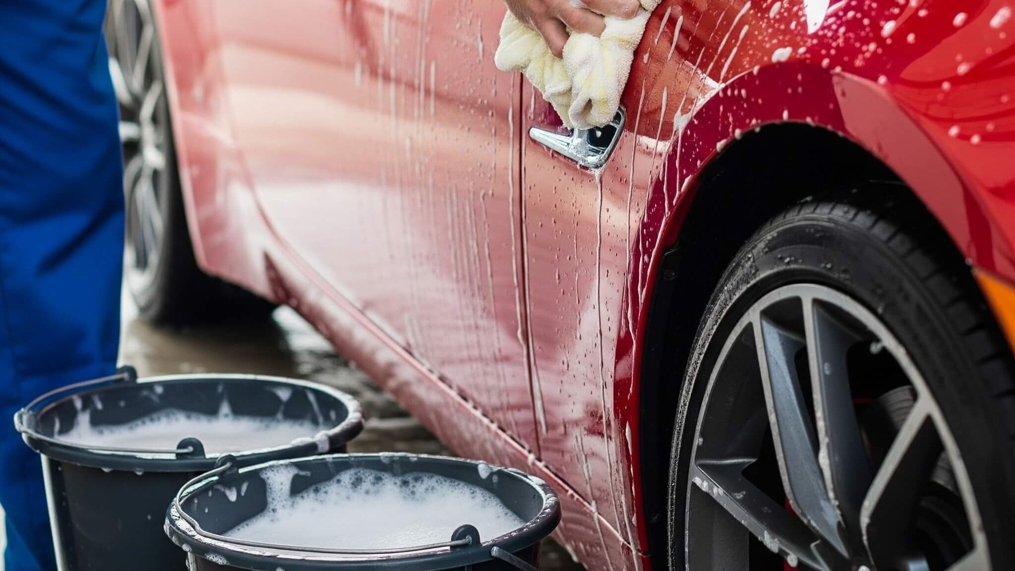Man in blue overalls hand-washing a red sports car using the two-bucket method. He is scrubbing the car's side panel with a cream-colored wash mitt, with suds and water dripping down the vehicle.