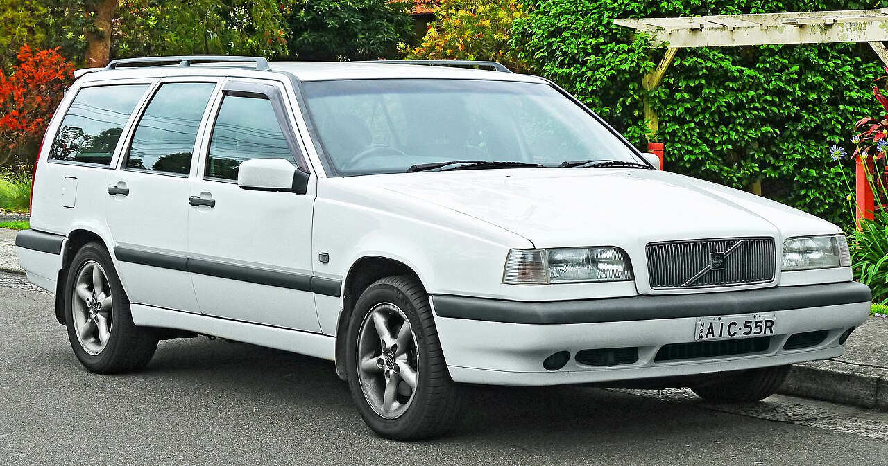 A white volvo estate from 1997 parked on a road next to green trees and leaves. The plate reads AIC - 55R. The car is boxy, so the engine will work harder and consume more fuel. 